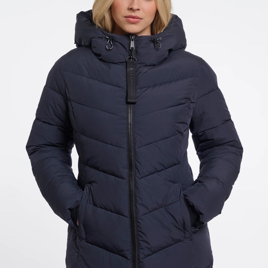 A woman wearing a long, dark navy puffer coat with a hood and front zipper, photographed against a plain light gray studio background.