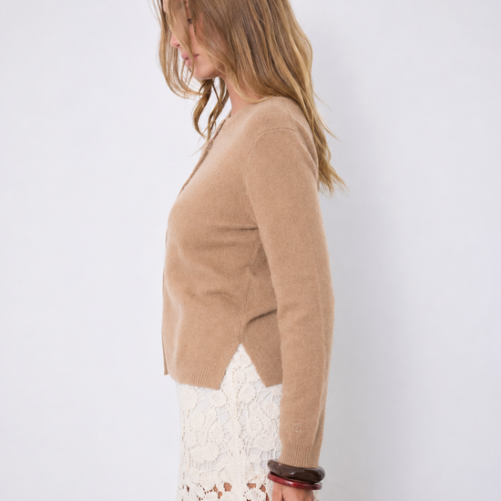 A woman shown in side profile wearing a cropped, long-sleeve knit cardigan in a warm camel tan color over a white lace skirt, set against a plain light gray studio background.
