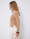 A woman shown in side profile wearing a cropped, long-sleeve knit cardigan in a warm camel tan color over a white lace skirt, set against a plain light gray studio background.