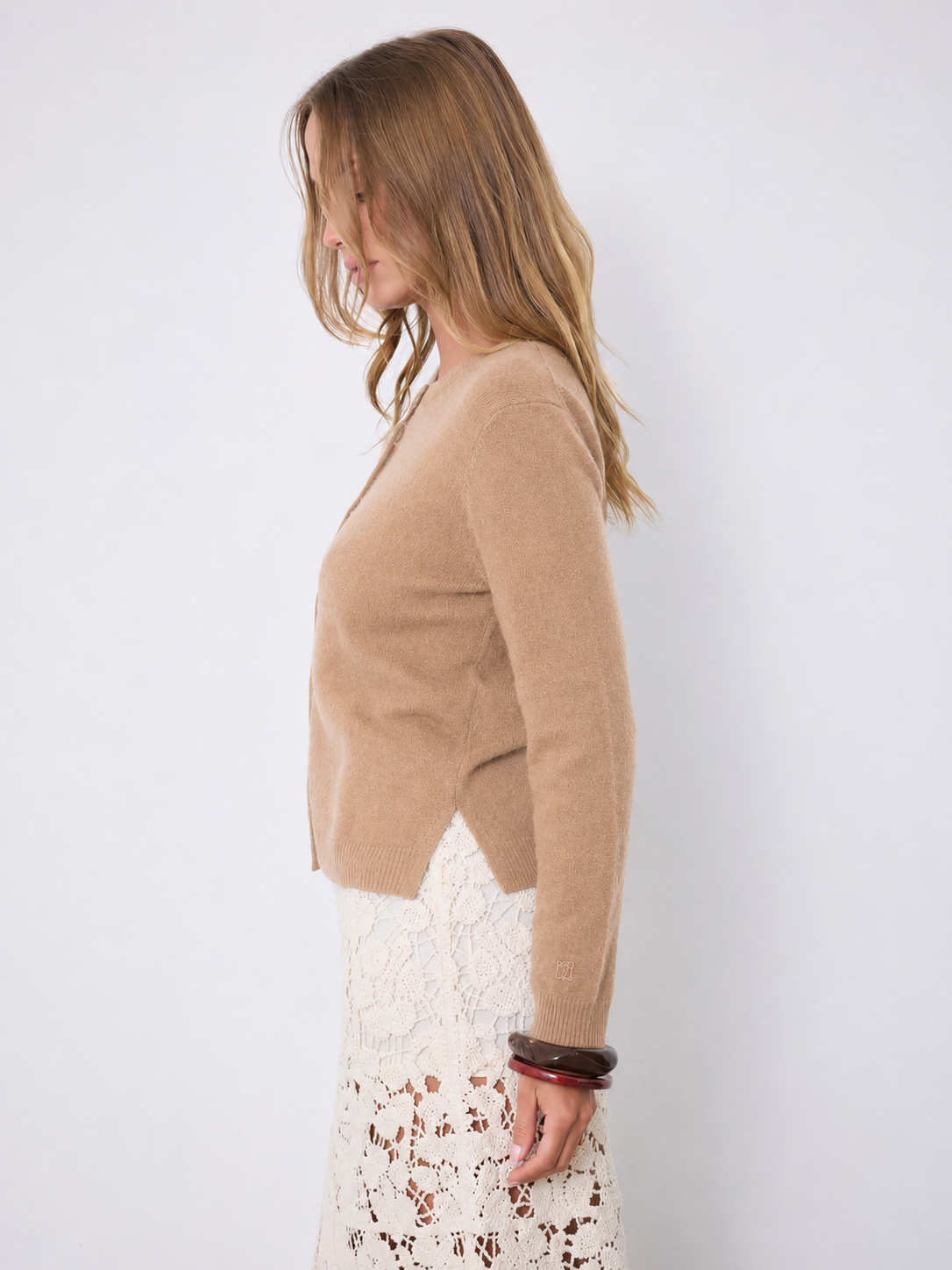 A woman shown in side profile wearing a cropped, long-sleeve knit cardigan in a warm camel tan color over a white lace skirt, set against a plain light gray studio background.