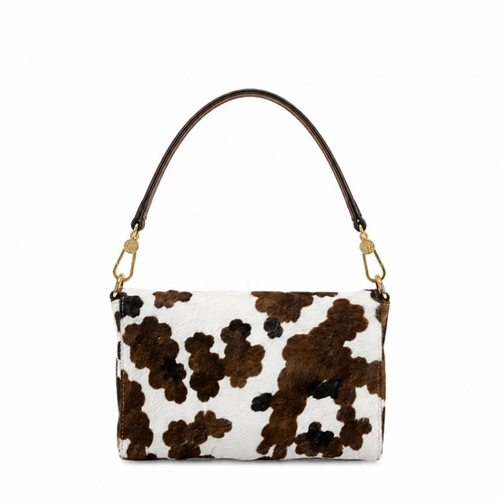 A small shoulder bag with a brown and black cow print pattern on a white background, featuring a dark brown leather shoulder strap and gold-tone hardware.