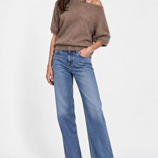 A woman modeling a brown off-the-shoulder knit sweater and wide-leg blue jeans against a plain light gray studio background.