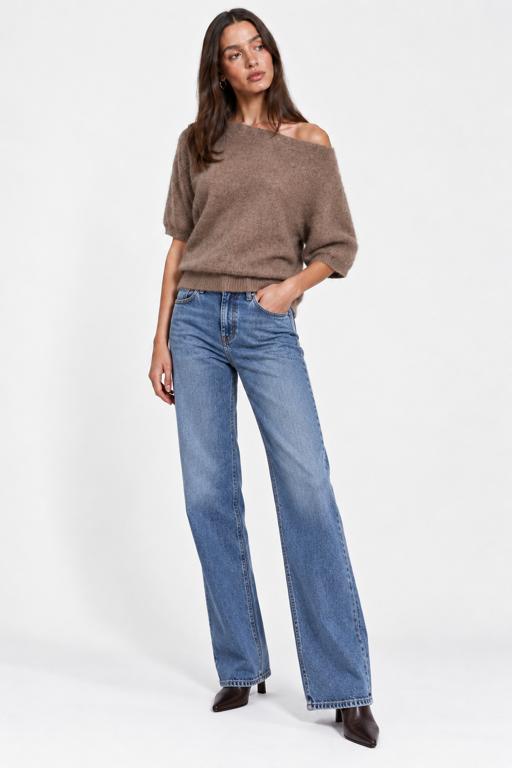 A woman modeling a brown off-the-shoulder knit sweater and wide-leg blue jeans against a plain light gray studio background.