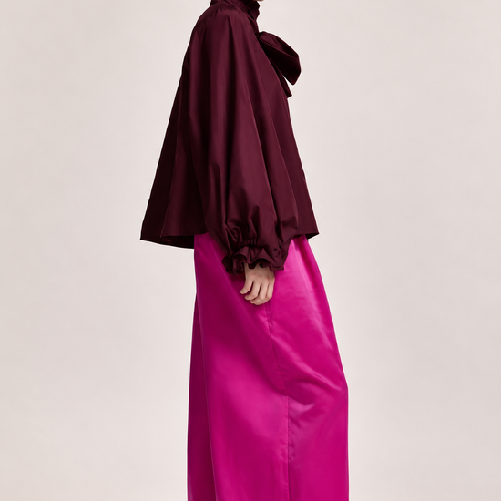 A model wearing a dark burgundy cape-style blouse with a high ruffled collar and pleated sleeves, paired with voluminous bright pink wide-leg trousers, against a solid light beige studio background.