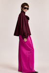 A model wearing a dark burgundy cape-style blouse with a high ruffled collar and pleated sleeves, paired with voluminous bright pink wide-leg trousers, against a solid light beige studio background.