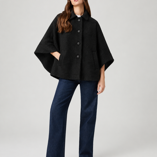 model wearing black cape style coat with blue denim jeans and black boots on a plain white background