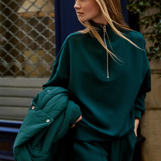 A woman wearing an oversized dark teal green half-zip sweatshirt and matching sweatpants, holding a matching green puffer jacket against a city storefront background.