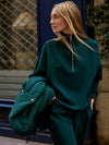 A woman wearing an oversized dark teal green half-zip sweatshirt and matching sweatpants, holding a matching green puffer jacket against a city storefront background.