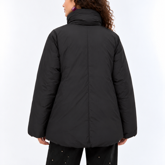 A woman wearing a black oversized puffer jacket with a high collar and front zipper, paired with black pants, photographed against a plain light gray studio background.