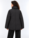 A woman wearing a black oversized puffer jacket with a high collar and front zipper, paired with black pants, photographed against a plain light gray studio background.