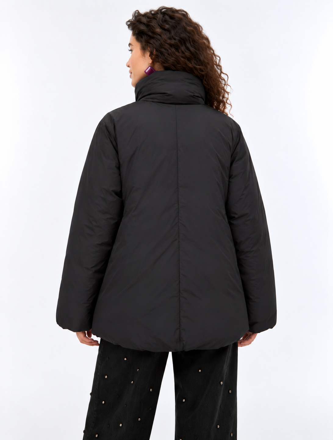 A woman wearing a black oversized puffer jacket with a high collar and front zipper, paired with black pants, photographed against a plain light gray studio background.