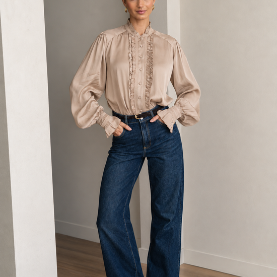 A woman stands in a minimalist room with light gray walls and wood floors, wearing a beige long-sleeve blouse with ruffle detailing down the front and dark blue wide-leg jeans.