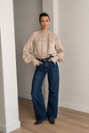 A woman stands in a minimalist room with light gray walls and wood floors, wearing a beige long-sleeve blouse with ruffle detailing down the front and dark blue wide-leg jeans.