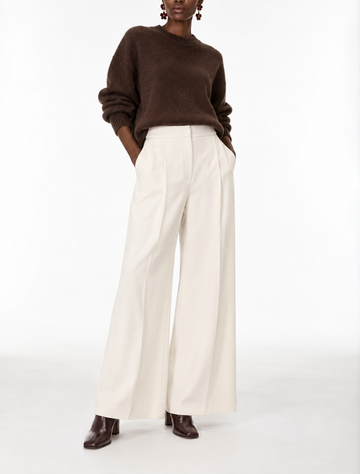A model wearing a brown knit sweater and high-waisted off-white wide-leg trousers, paired with dark brown leather shoes, against a plain white studio background.