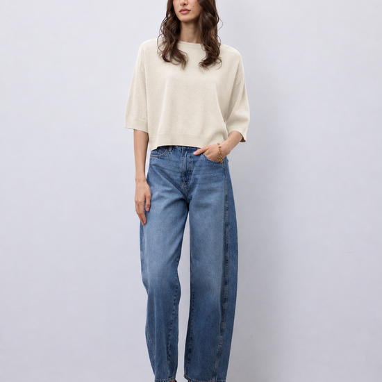 A woman modeling a cream short-sleeve knit top and wide-leg blue jeans with snakeskin heels, standing against a light gray studio background.
