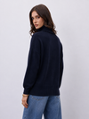 A woman wearing a dark navy blue ribbed knit sweater with a high mock neck, shown from a three-quarter back view against a light gray studio background.