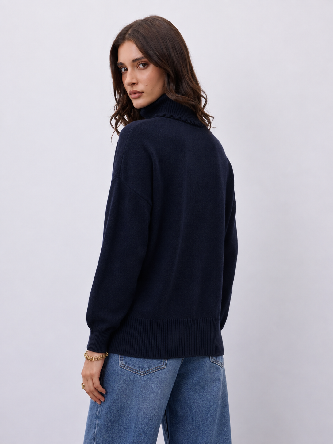 A woman wearing a dark navy blue ribbed knit sweater with a high mock neck, shown from a three-quarter back view against a light gray studio background.