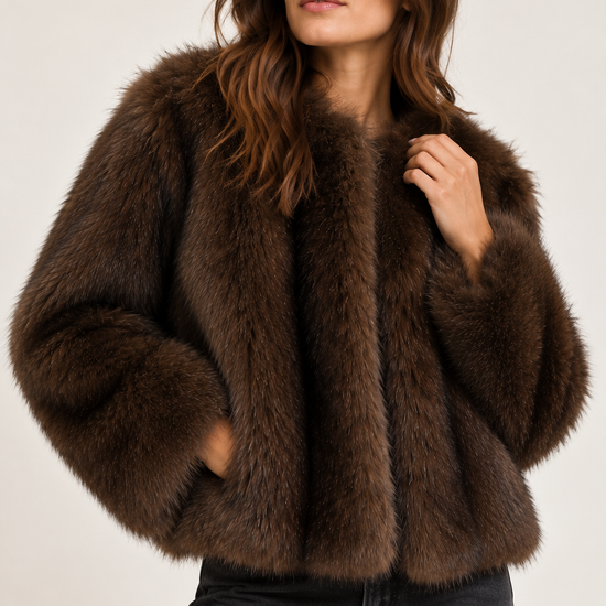 A woman wearing a dark brown shaggy faux fur cropped jacket and black jeans, posing against a light gray studio background.