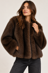 A woman wearing a dark brown shaggy faux fur cropped jacket and black jeans, posing against a light gray studio background.