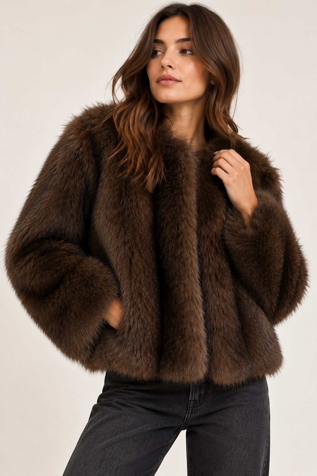 A woman wearing a dark brown shaggy faux fur cropped jacket and black jeans, posing against a light gray studio background.