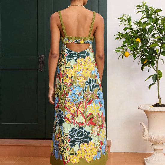 Woman wearing a colorful floral dress standing in front of a green door with a plant to her right from Alemais