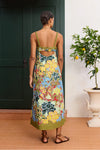 Woman wearing a colorful floral dress standing in front of a green door with a plant to her right from Alemais