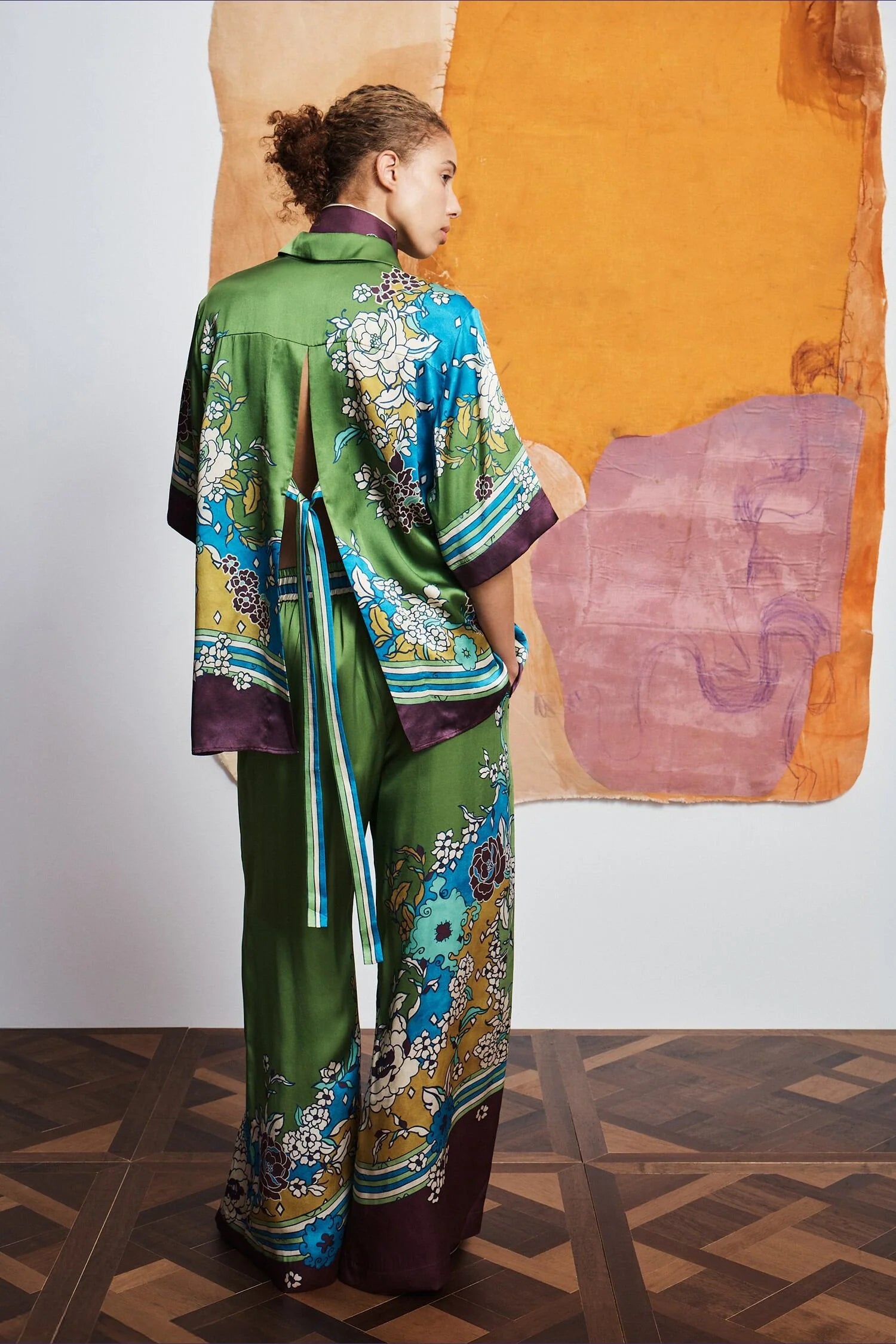 Woman wearing a colorful outfit with a patterned shirt and pants by Alemais against an abstract wall rear view