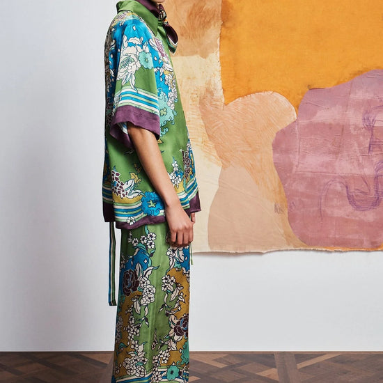 Woman wearing a colorful outfit with a patterned shirt and pants by Alemais against an abstract wall side view