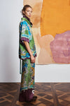 Woman wearing a colorful outfit with a patterned shirt and pants by Alemais against an abstract wall side view