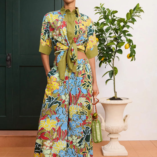 Woman wearing a colorful floral outfit standing in a room with a plant and door.