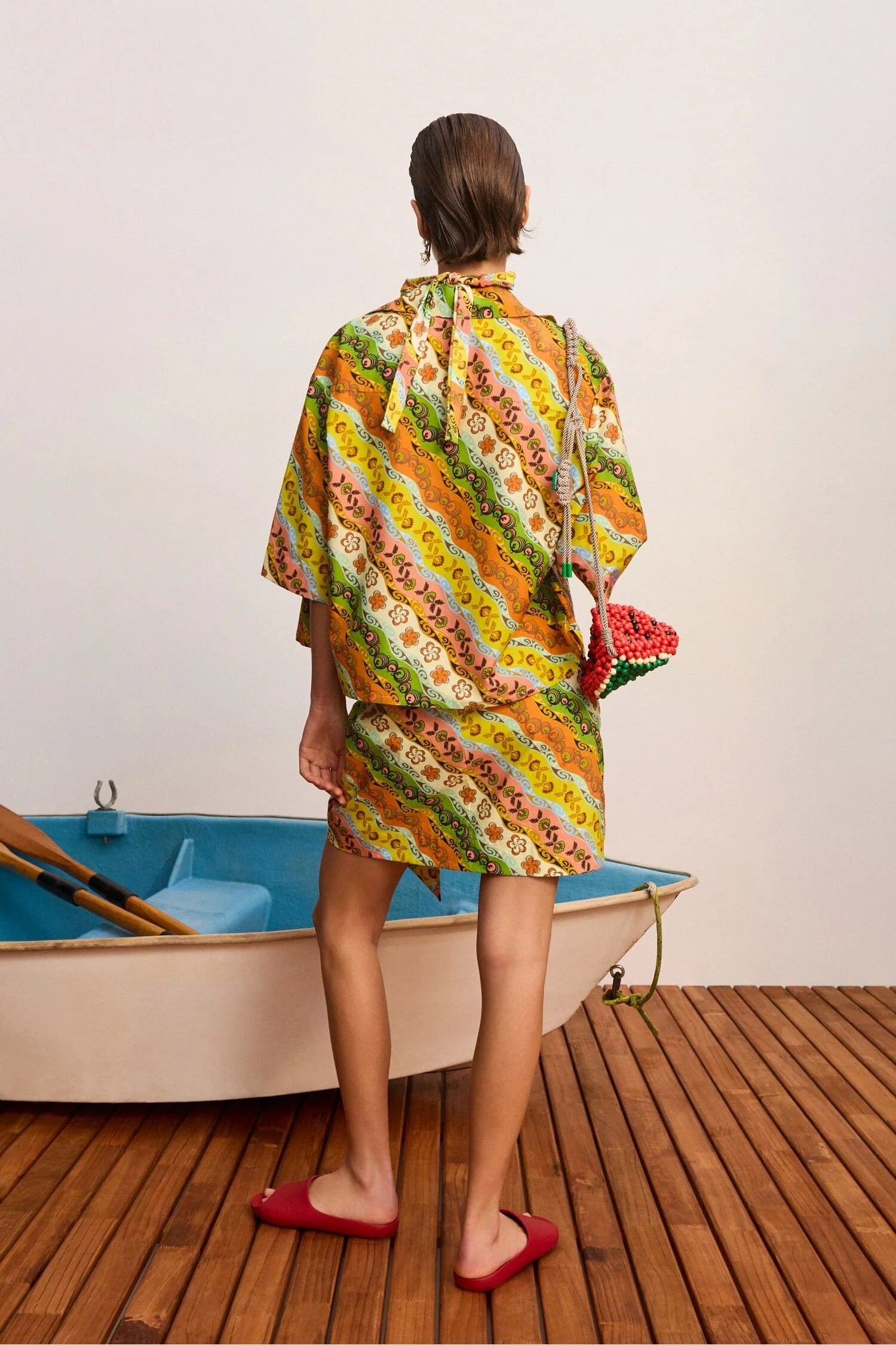 Woman in a colorful patterned dress standing next to a small boat on a wooden deck.