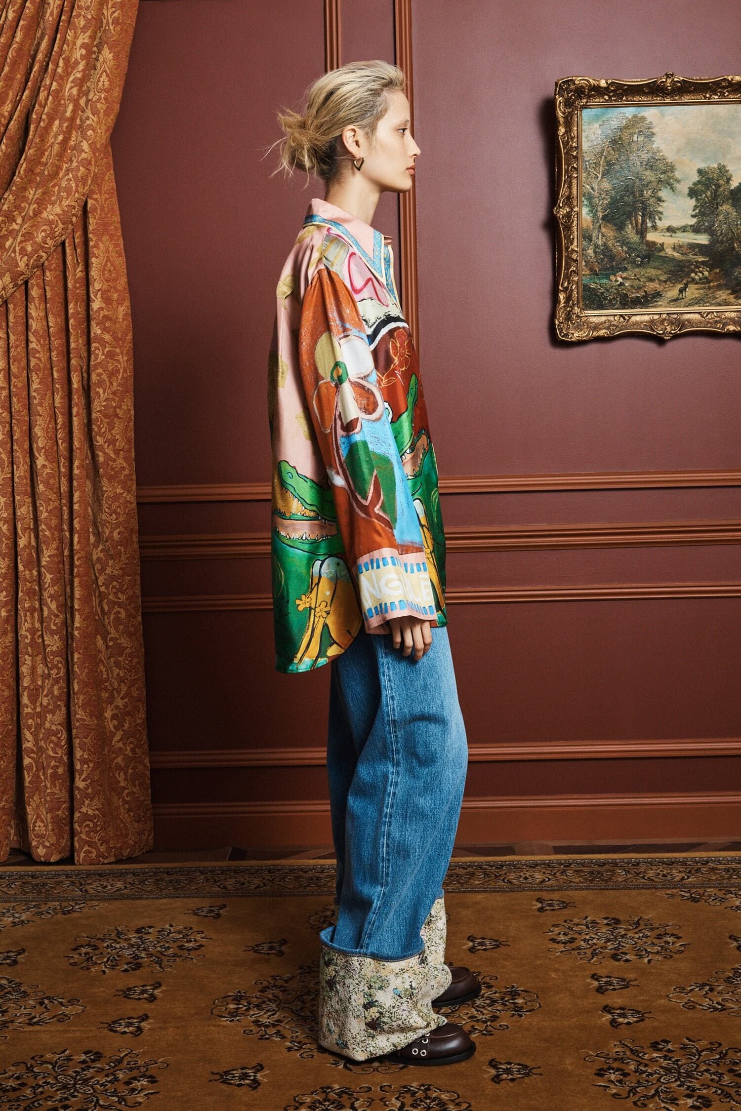 Person wearing a colorful shirt by Alemais and blue jeans in an elegant room with a painting on the wall side view