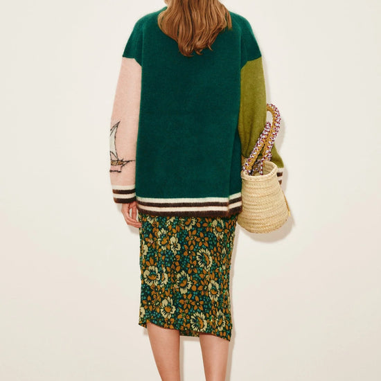 Rear view of Woman wearing a green and brown color-blocked cardigan with a floral skirt, holding a woven bag from Alemais