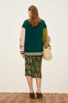 Rear view of Woman wearing a green and brown color-blocked cardigan with a floral skirt, holding a woven bag from Alemais