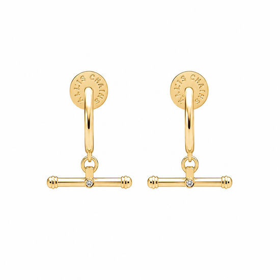 A pair of gold huggie hoop earrings with a dangling barbell charm and a small clear rhinestone in the center, shown on a plain white background.