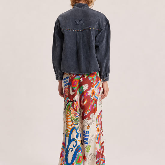 A model shown from the back wearing a dark denim jacket with silver studs and a long, wide skirt featuring a vibrant multicolor abstract print, set against a plain light beige studio background.