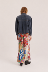 A model shown from the back wearing a dark denim jacket with silver studs and a long, wide skirt featuring a vibrant multicolor abstract print, set against a plain light beige studio background.