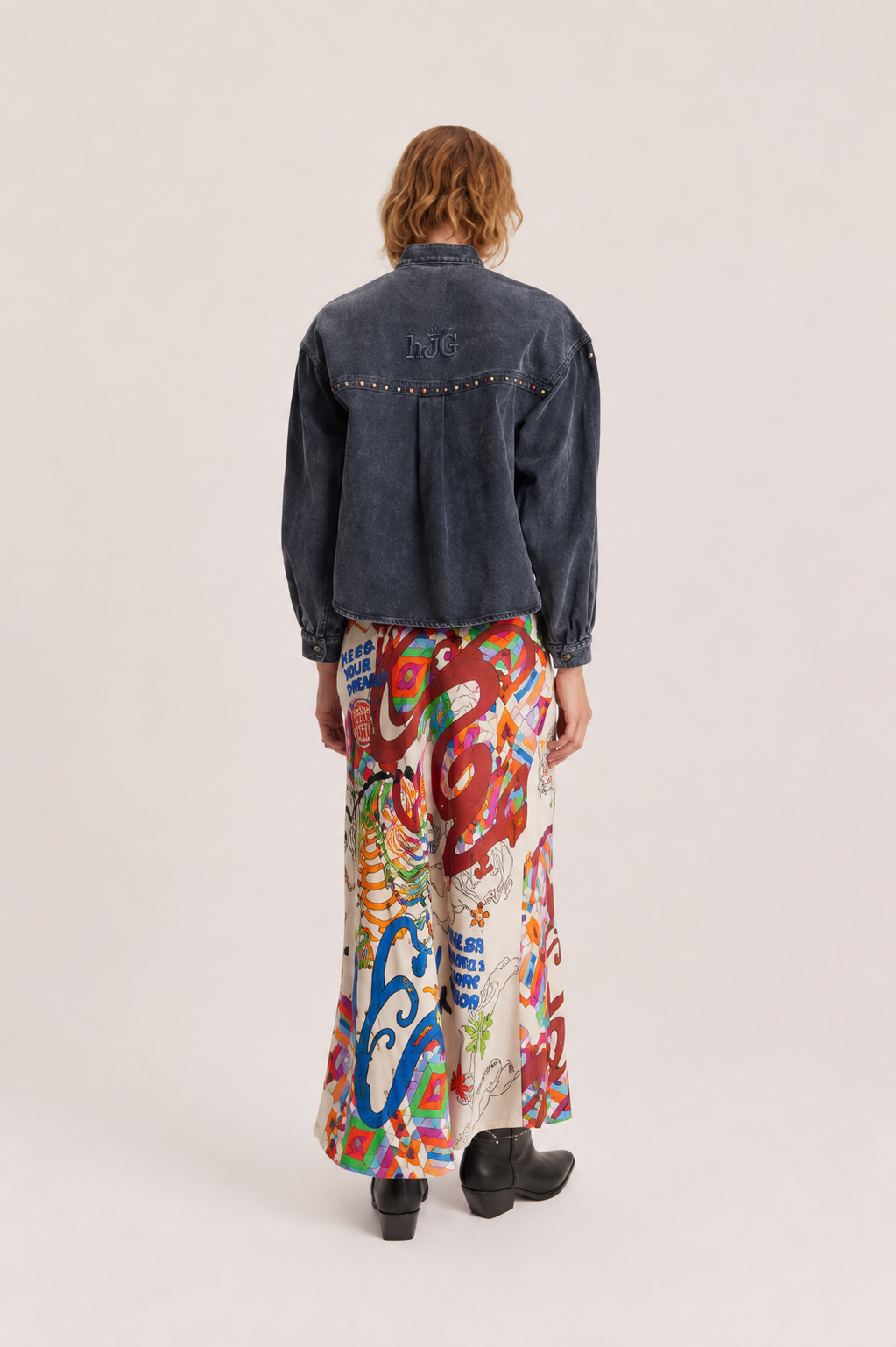 A model shown from the back wearing a dark denim jacket with silver studs and a long, wide skirt featuring a vibrant multicolor abstract print, set against a plain light beige studio background.