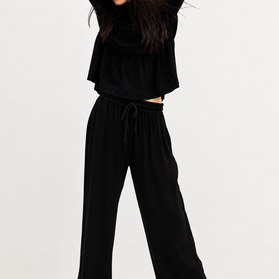 A model wearing a black long-sleeve cropped top, matching black wide-leg drawstring pants, a black baseball cap, and black shoes, posing against a plain white studio background.