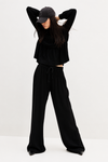 A model wearing a black long-sleeve cropped top, matching black wide-leg drawstring pants, a black baseball cap, and black shoes, posing against a plain white studio background.