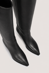 Black leather pointed-toe knee-high boots shown from a high-angle close-up on a light gray background.