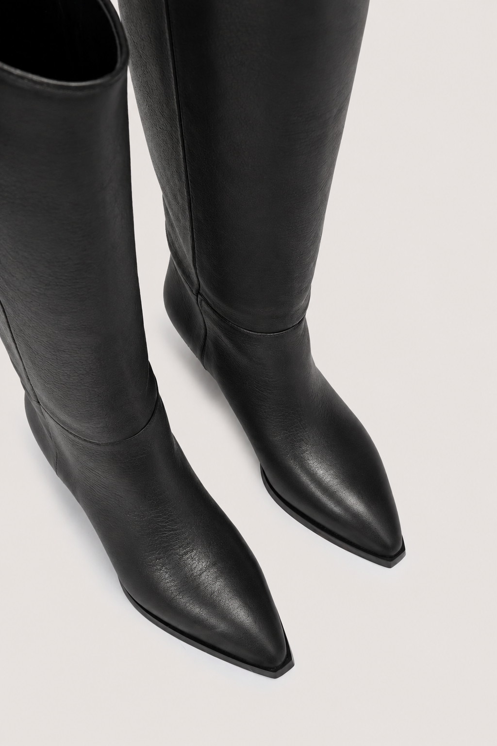 Black leather pointed-toe knee-high boots shown from a high-angle close-up on a light gray background.