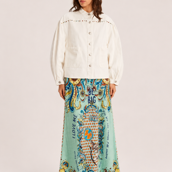 A model stands against a light beige studio background wearing a cream-colored cropped jacket with puff sleeves and a long, flowing mint green skirt featuring a colorful, ornate paisley and floral pattern with text.