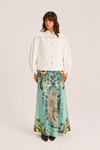 A model stands against a light beige studio background wearing a cream-colored cropped jacket with puff sleeves and a long, flowing mint green skirt featuring a colorful, ornate paisley and floral pattern with text.