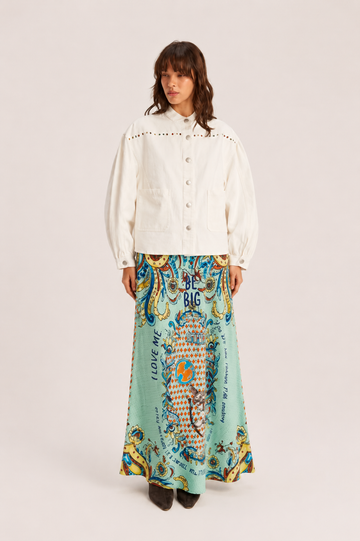 A model stands against a light beige studio background wearing a cream-colored cropped jacket with puff sleeves and a long, flowing mint green skirt featuring a colorful, ornate paisley and floral pattern with text.
