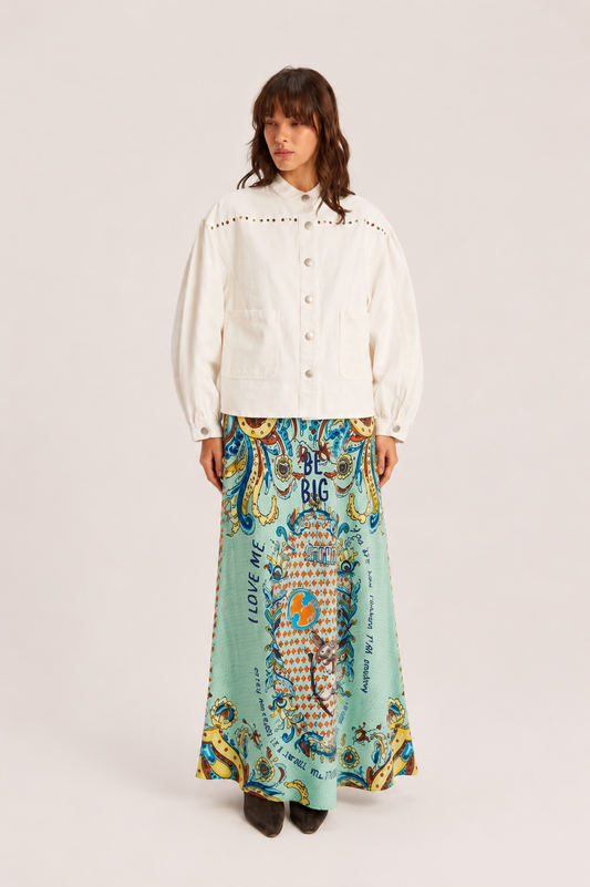 A model stands against a light beige studio background wearing a cream-colored cropped jacket with puff sleeves and a long, flowing mint green skirt featuring a colorful, ornate paisley and floral pattern with text.