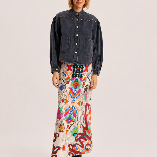 A model stands against a light gray studio background wearing a dark denim cropped jacket with decorative stitching, a long white maxi skirt with a vibrant multicolor abstract print, and black pointed-toe boots.