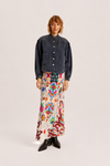 A model stands against a light gray studio background wearing a dark denim cropped jacket with decorative stitching, a long white maxi skirt with a vibrant multicolor abstract print, and black pointed-toe boots.