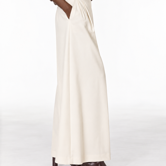 A person wearing a cream wide-leg skirt with pleated detailing at the waist, styled with a brown sweater and dark brown heeled boots against a clean white studio background.