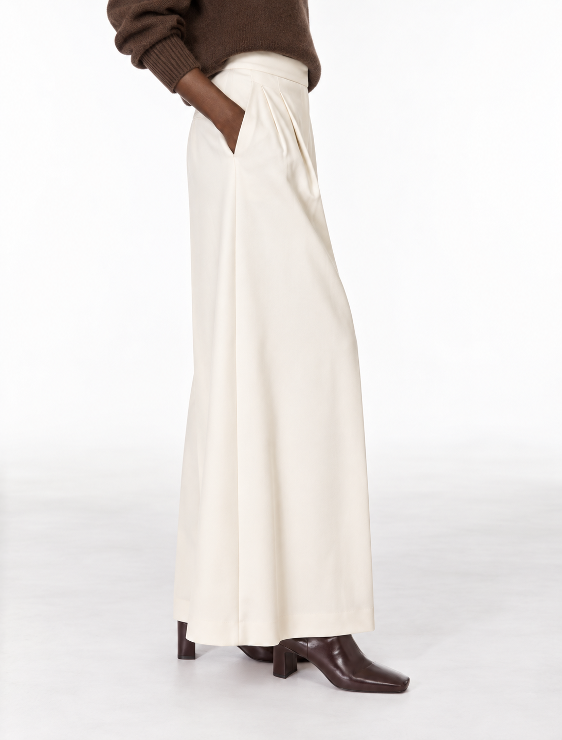 A person wearing a cream wide-leg skirt with pleated detailing at the waist, styled with a brown sweater and dark brown heeled boots against a clean white studio background.